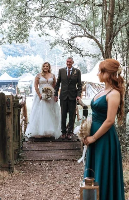 Bride and groom standing on a bridge outdoors with a bridesmaid holding a lantern in the foreground; tent structures visible in the background.