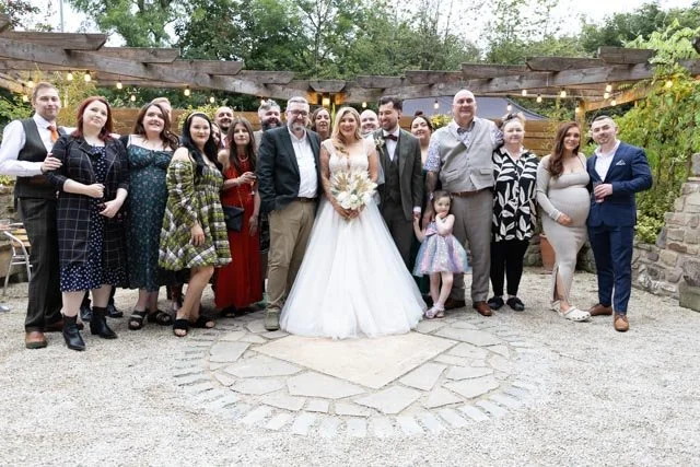 Large group of people posing for a photo at an outdoor wedding ceremony, with a bride and groom in formal attire at the center, surrounded by family and friends under a rustic pergola with string lights.