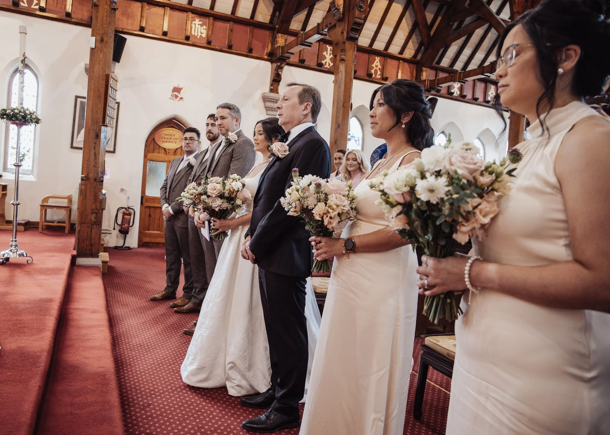 Wedding ceremony inside a church with the bride, groom, bridesmaids, and groomsmen standing at the altar, holding bouquets of flowers.
