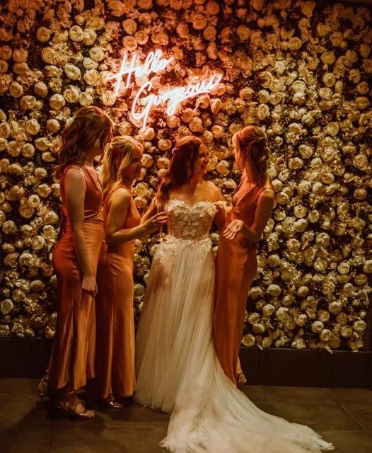 Bride and bridesmaids in formal dresses standing in front of a flower wall with a neon sign reading 'Hello Gorgeous.'