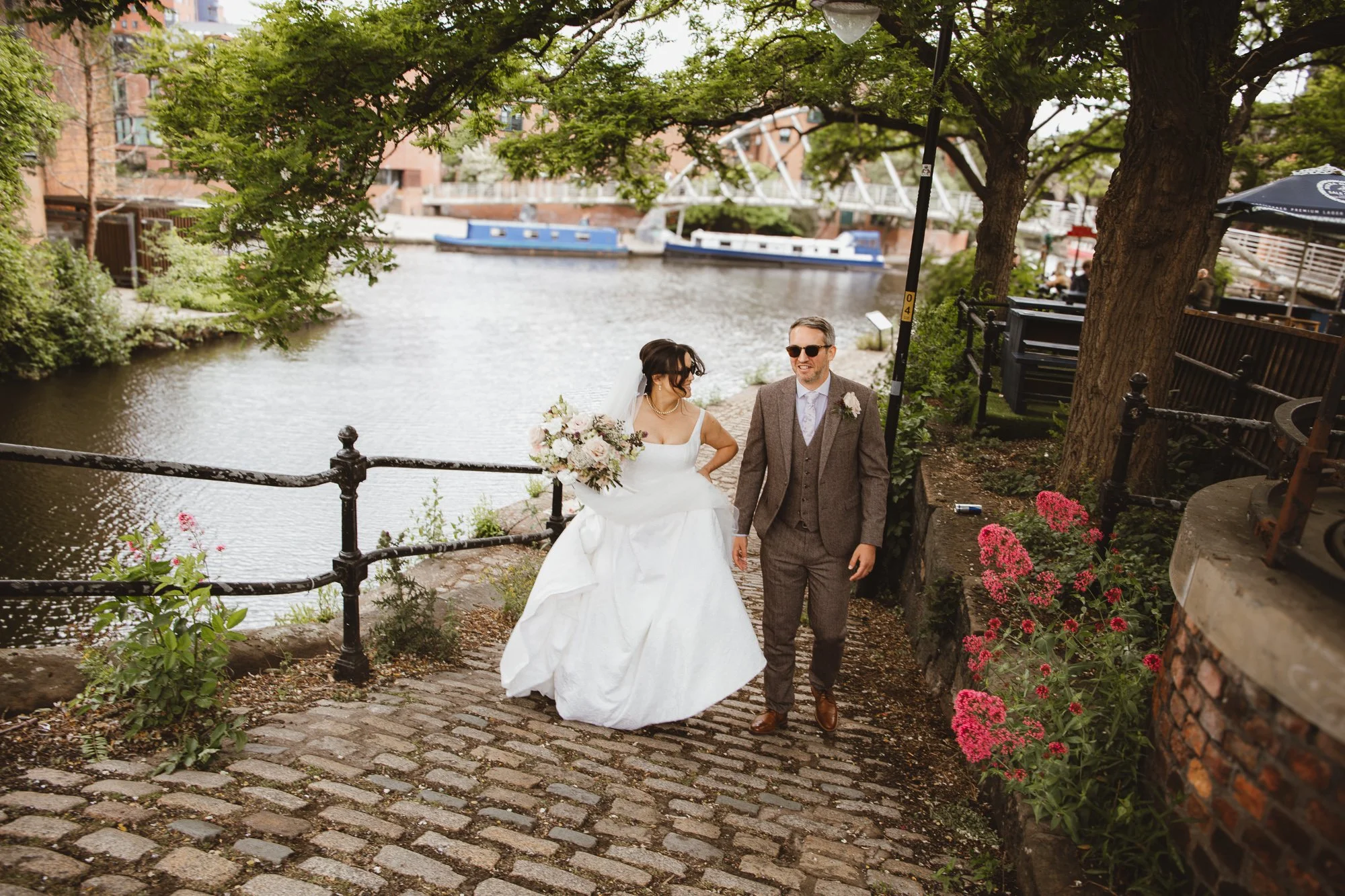 Bride and groom walking along a cobblestone path beside a river, surrounded by greenery and pink flowers, with boats in the background.