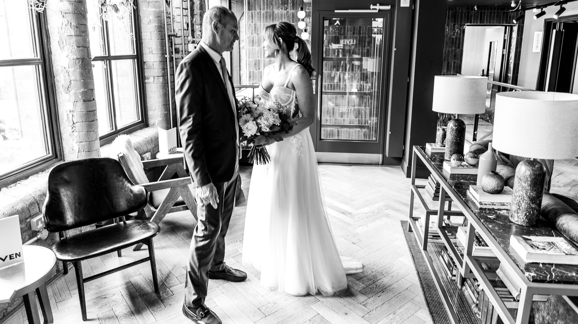 Black and white photo of a bride in a wedding dress holding a bouquet standing with an older man in a suit in a stylish interior with large windows and modern furnishings.