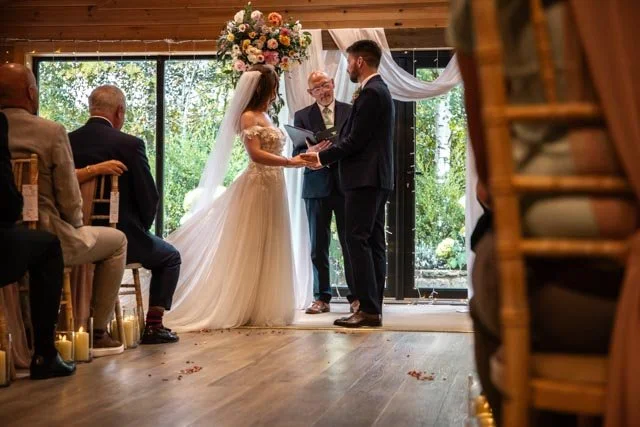 Indoor wedding ceremony with bride and groom holding hands, officiant standing between them under floral arch, surrounded by seated guests.