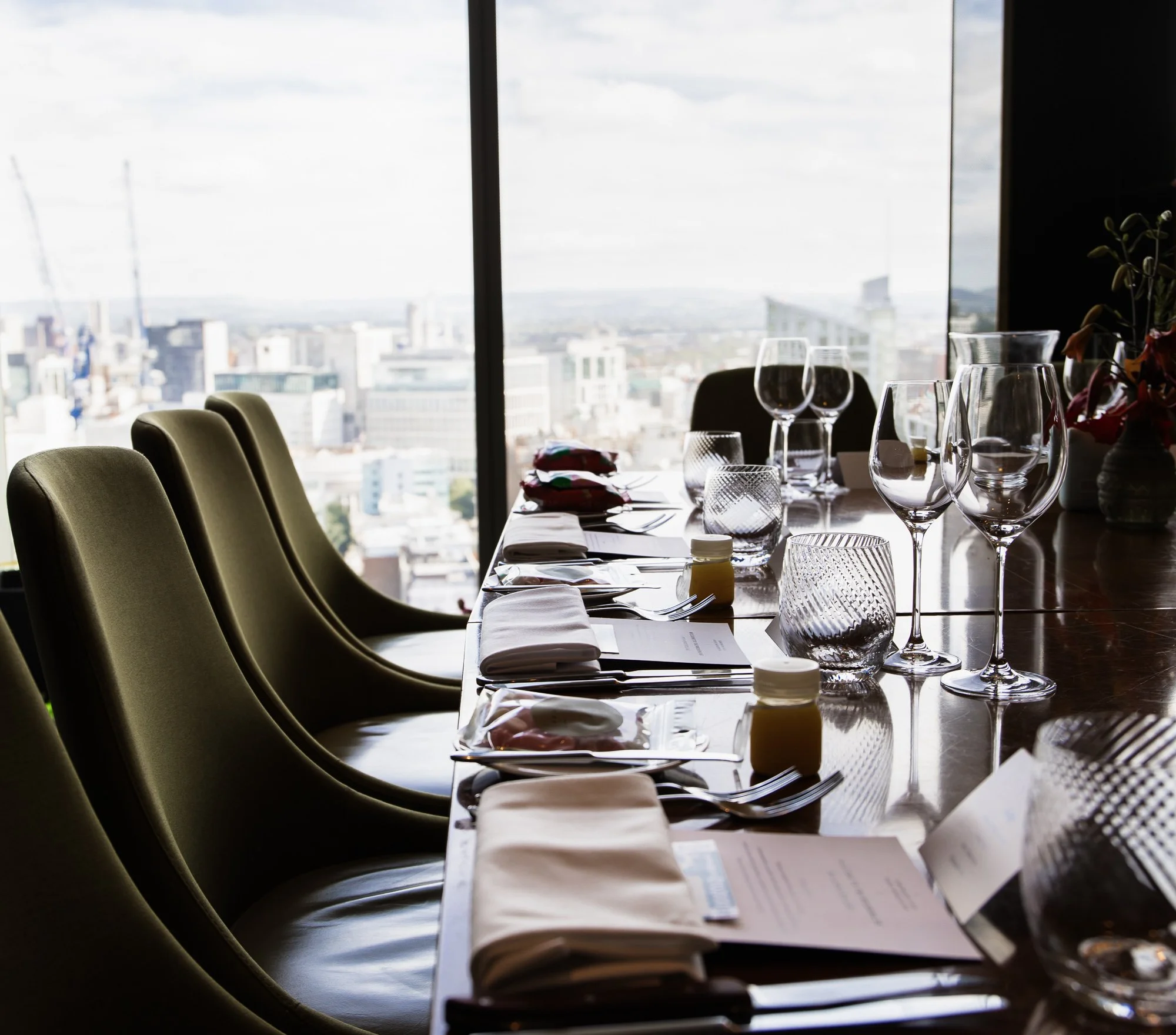 Elegant dining table set for a formal event with wine glasses, plates, and folded napkins, overlooking a city skyline through a large window.
