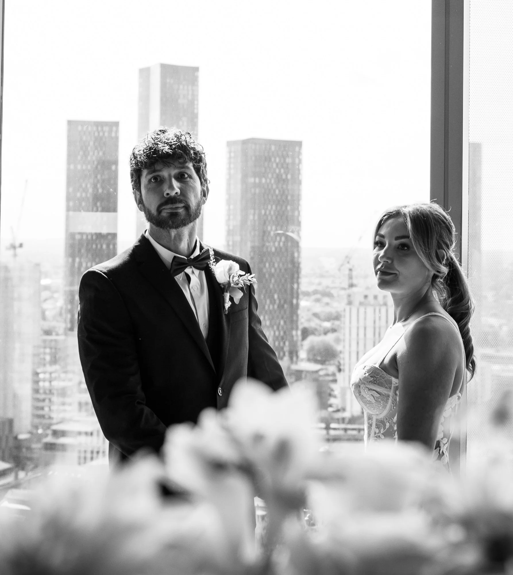 Black and white photo of a bride and groom standing indoors with city skyscrapers visible through a window behind them.