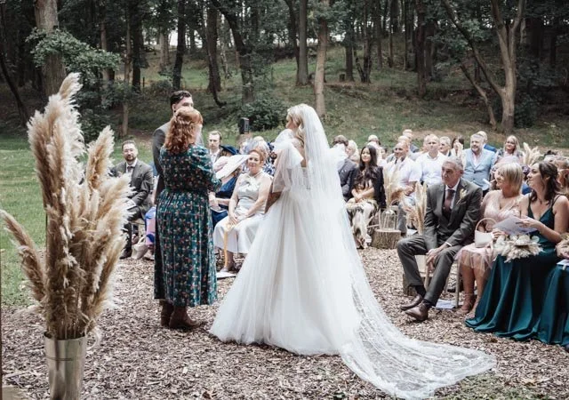 Outdoor wedding ceremony in a wooded area with a bride in a white gown and veil facing an officiant, guests seated on wooden benches, and pampas grass decorations.