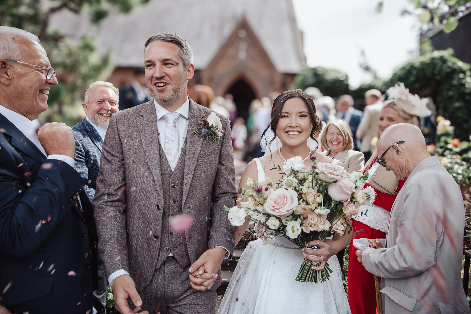 Bride and groom holding hands and smiling at their wedding reception, surrounded by family and friends outside a church, with flowers and confetti in the air.