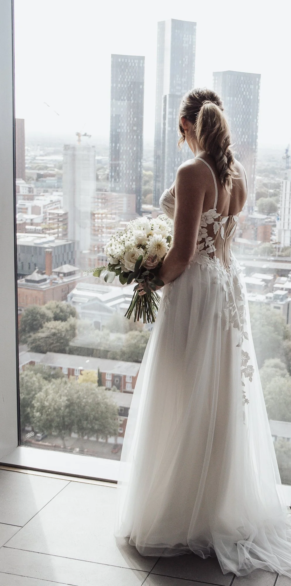 Bride in lace wedding dress holding bouquet, standing by window with city skyline view.