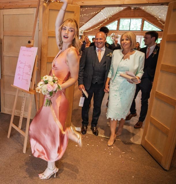 A woman in a pink dress joyfully holds a bouquet near a wedding entrance, with two older guests smiling and walking toward the event inside a decorated venue.