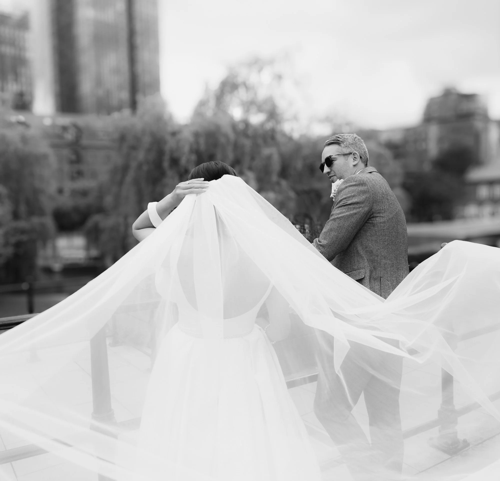 A black and white photo of a bride and groom on a bridge, with the bride’s veil covering her face, and the groom wearing sunglasses and a suit.