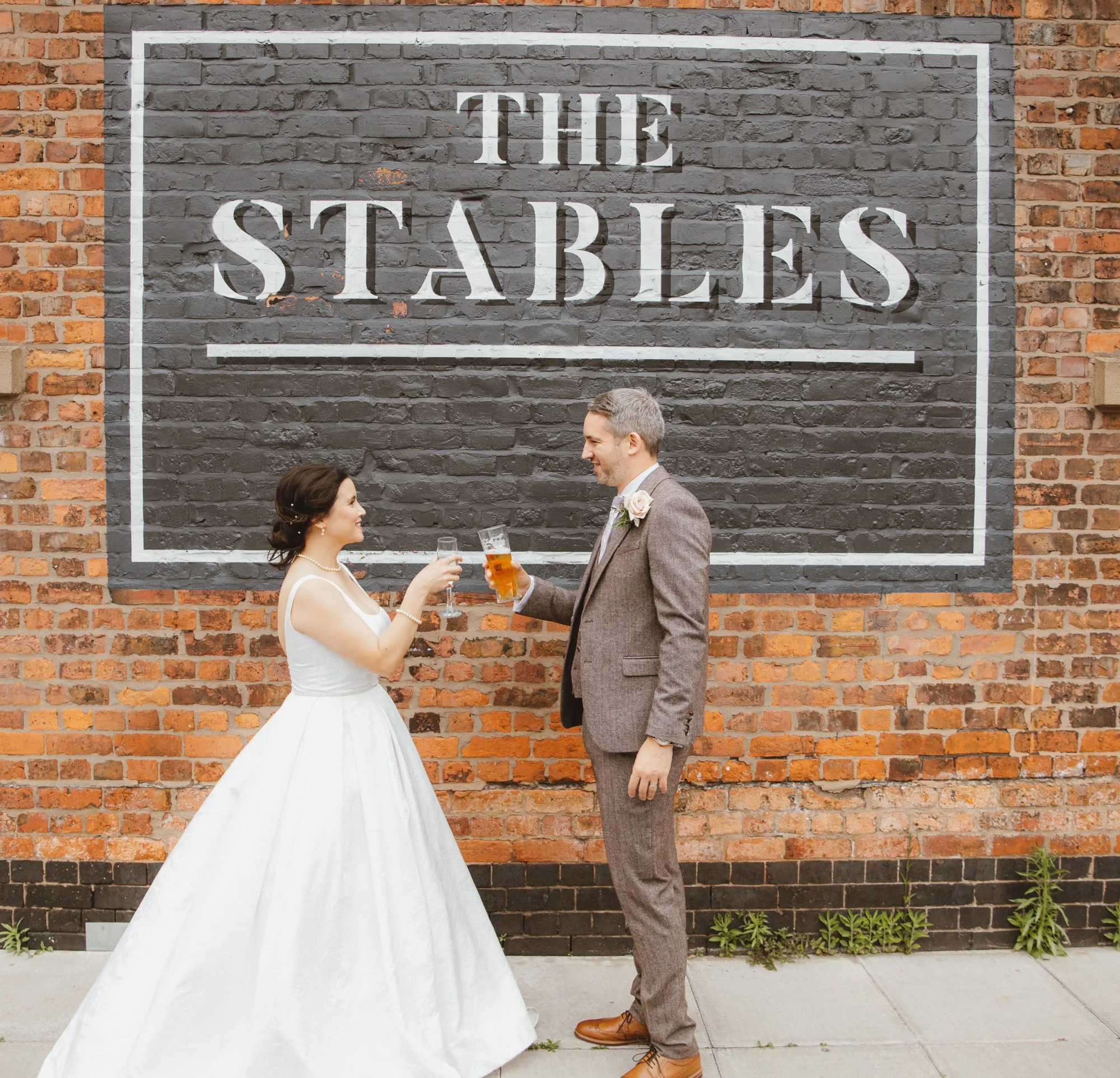 A bride and groom clinking glasses of beer in front of a brick wall with a large sign that reads 'THE STABLES'.