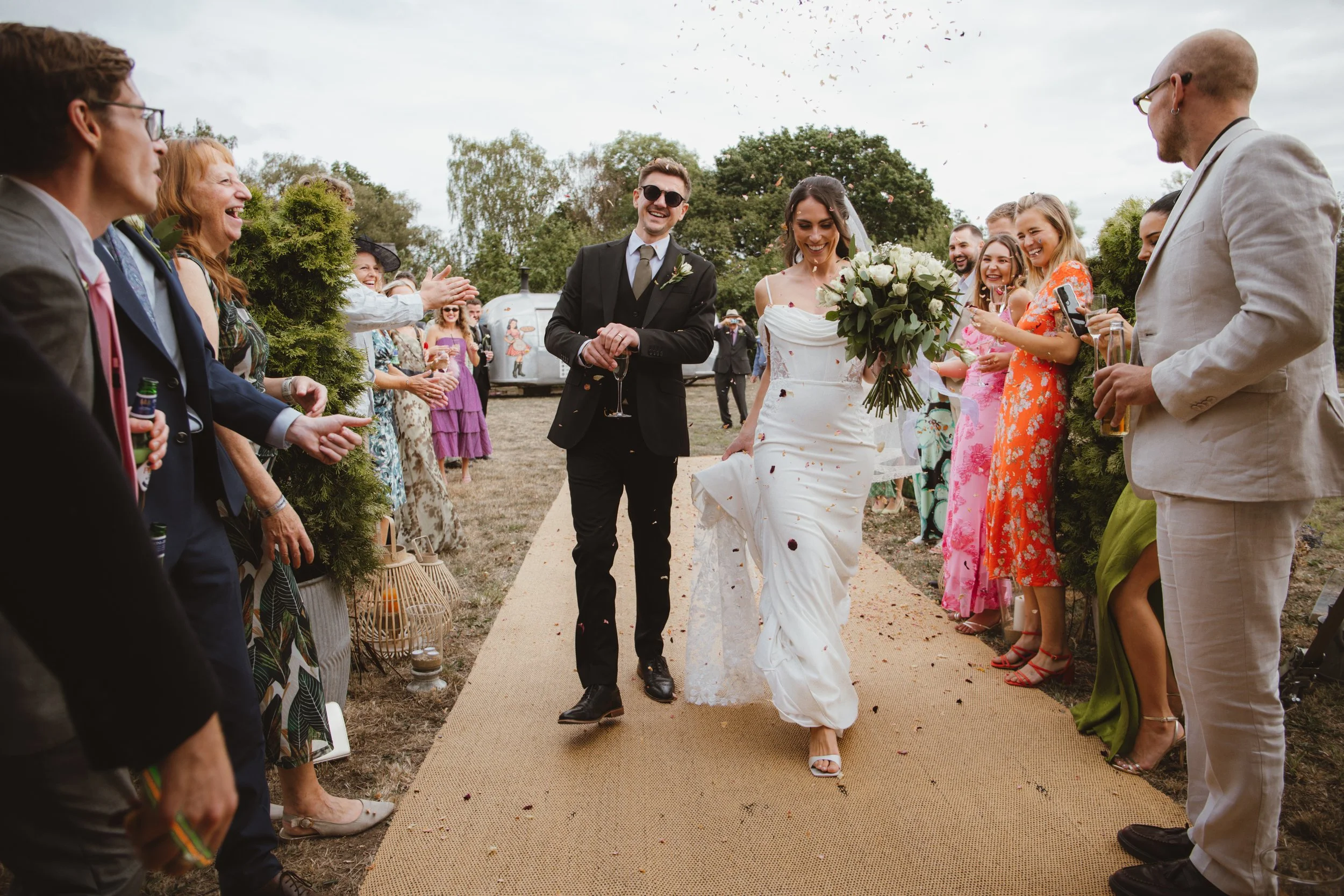 A bride and groom walk down an outdoor aisle, smiling, surrounded by joyful guests on both sides. The bride is holding a bouquet of white flowers, and the groom is in a black suit with sunglasses. Guests are clapping and celebrating, with some capturing the moment. Confetti is falling from above, and the scene is set in a grassy outdoor area with trees and a trailer in the background.