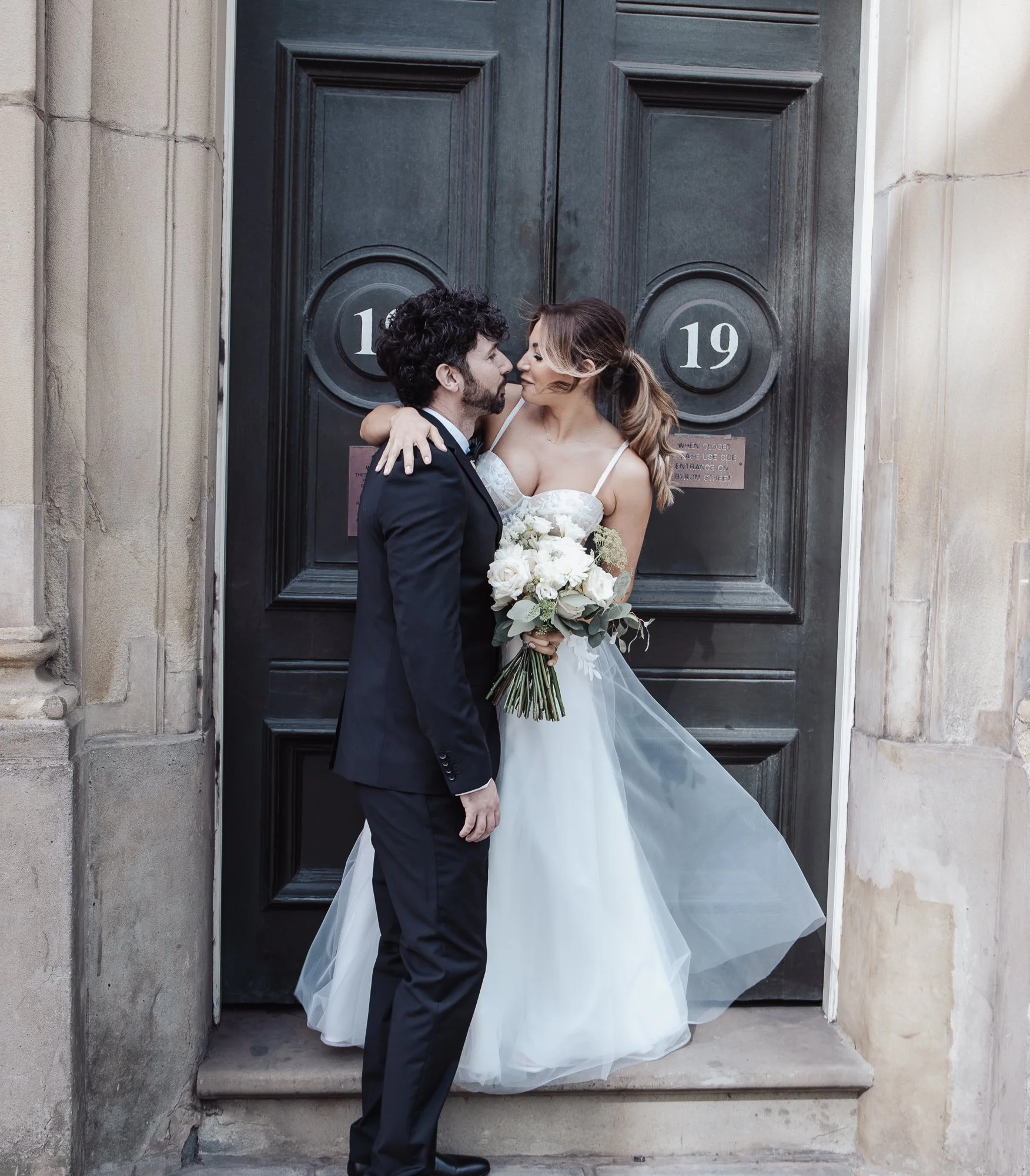 Bride and groom embracing in front of large wooden doors, bride holding bouquet of white flowers.