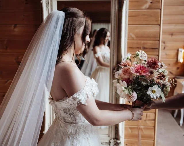 Bride holding a bouquet of flowers, standing in front of a mirror in a wooden room.