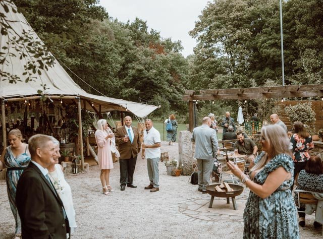 Outdoor event with people socializing near a tent, surrounded by trees and greenery, with wooden benches and tables.