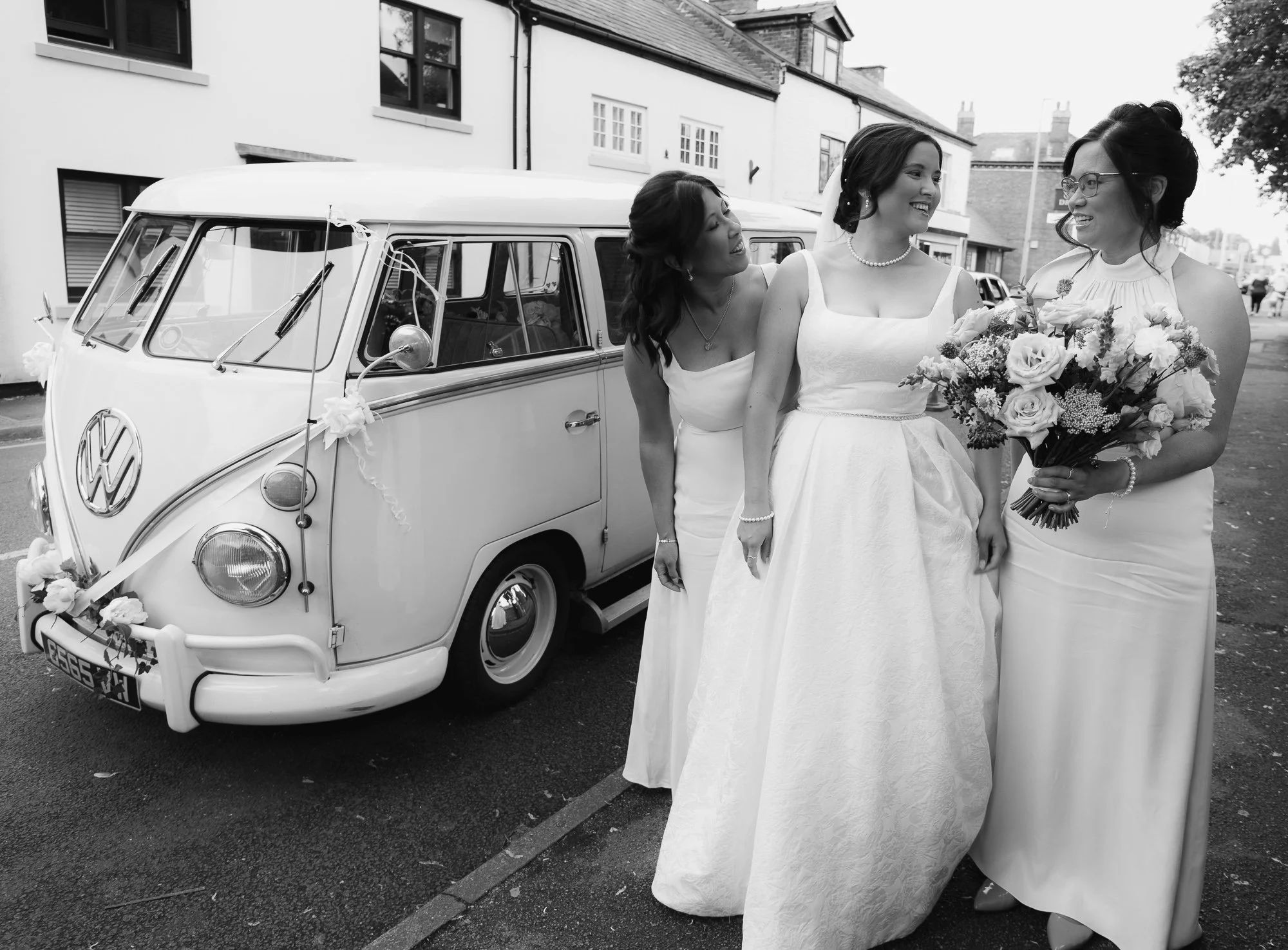 Black and white photo of four women in wedding dresses standing next to a vintage Volkswagen van decorated with flowers, on a street with houses in the background.