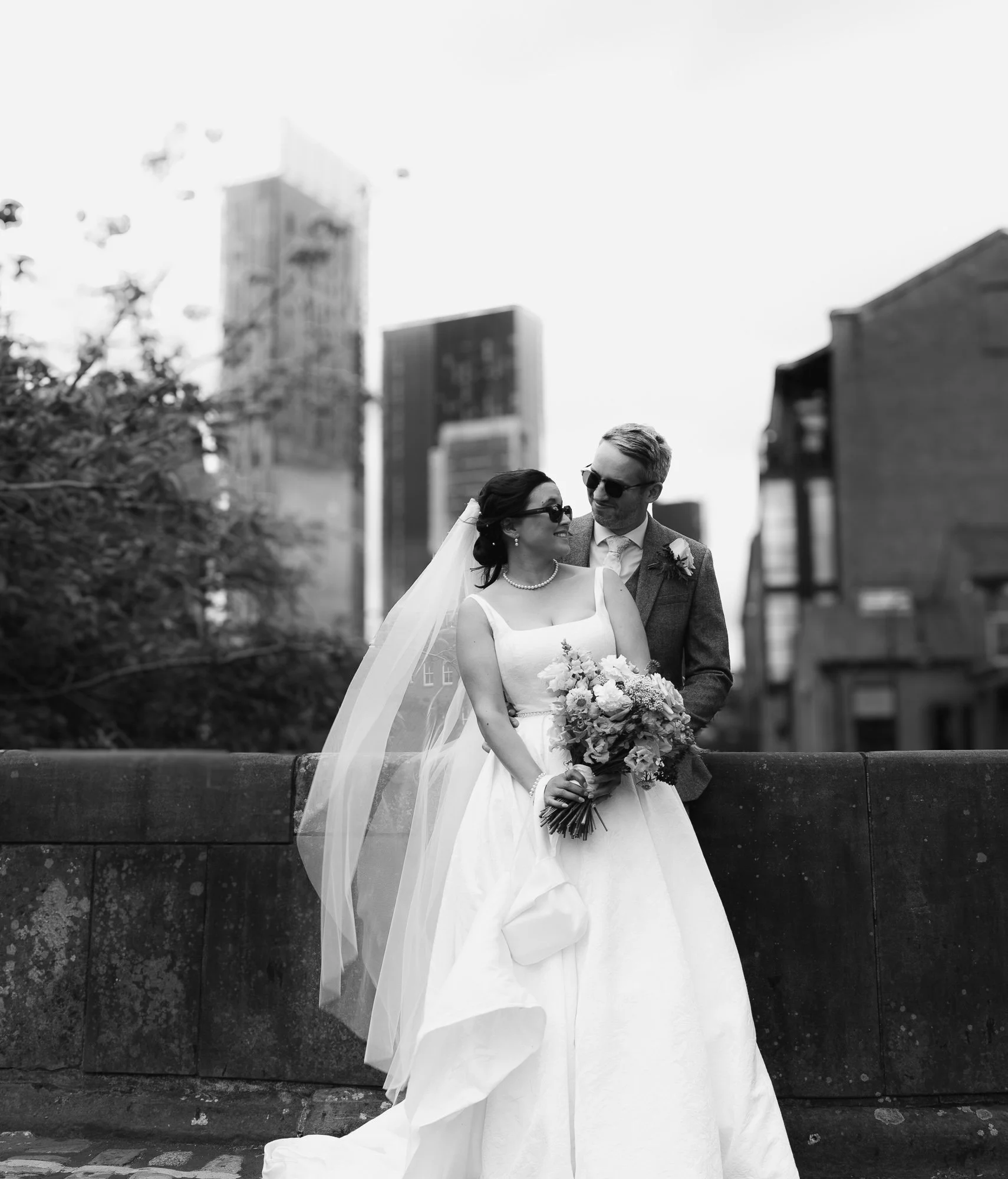 A black and white photo of a bride and groom standing close on a city bridge; the bride wears sunglasses, a pearl necklace, and a veil, holding a bouquet, while the groom wears sunglasses and a suit with a boutonniere, with city buildings in the background.