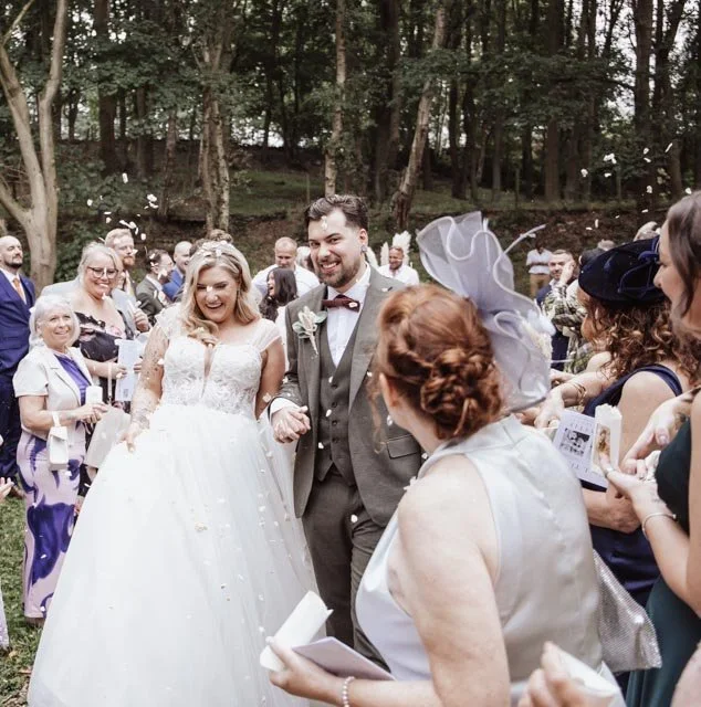 A bride and groom walking hand-in-hand at an outdoor wedding ceremony, surrounded by guests throwing confetti. The bride is in a white gown and the groom is in a suit. The setting is a wooded area, and guests are dressed in formal attire.