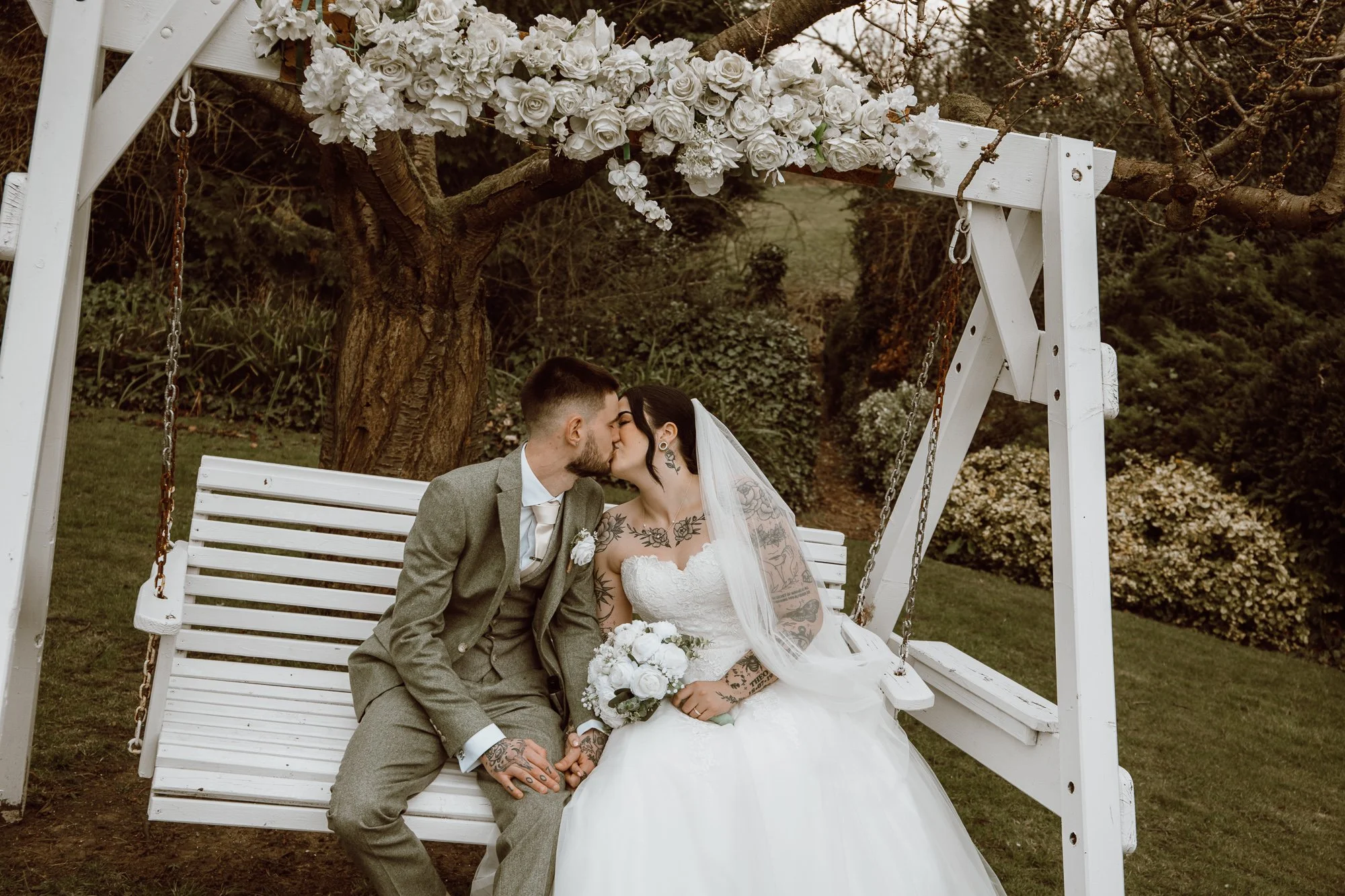 Bride and groom kissing on a white swing, surrounded by flowers, in a garden setting.