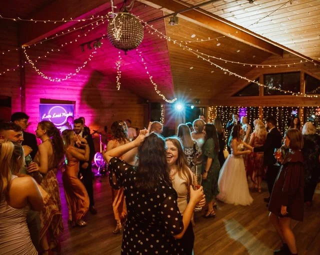 Group of people dancing at a lively indoor party with a disco ball, fairy lights, and a live band setup under purple and blue lighting.