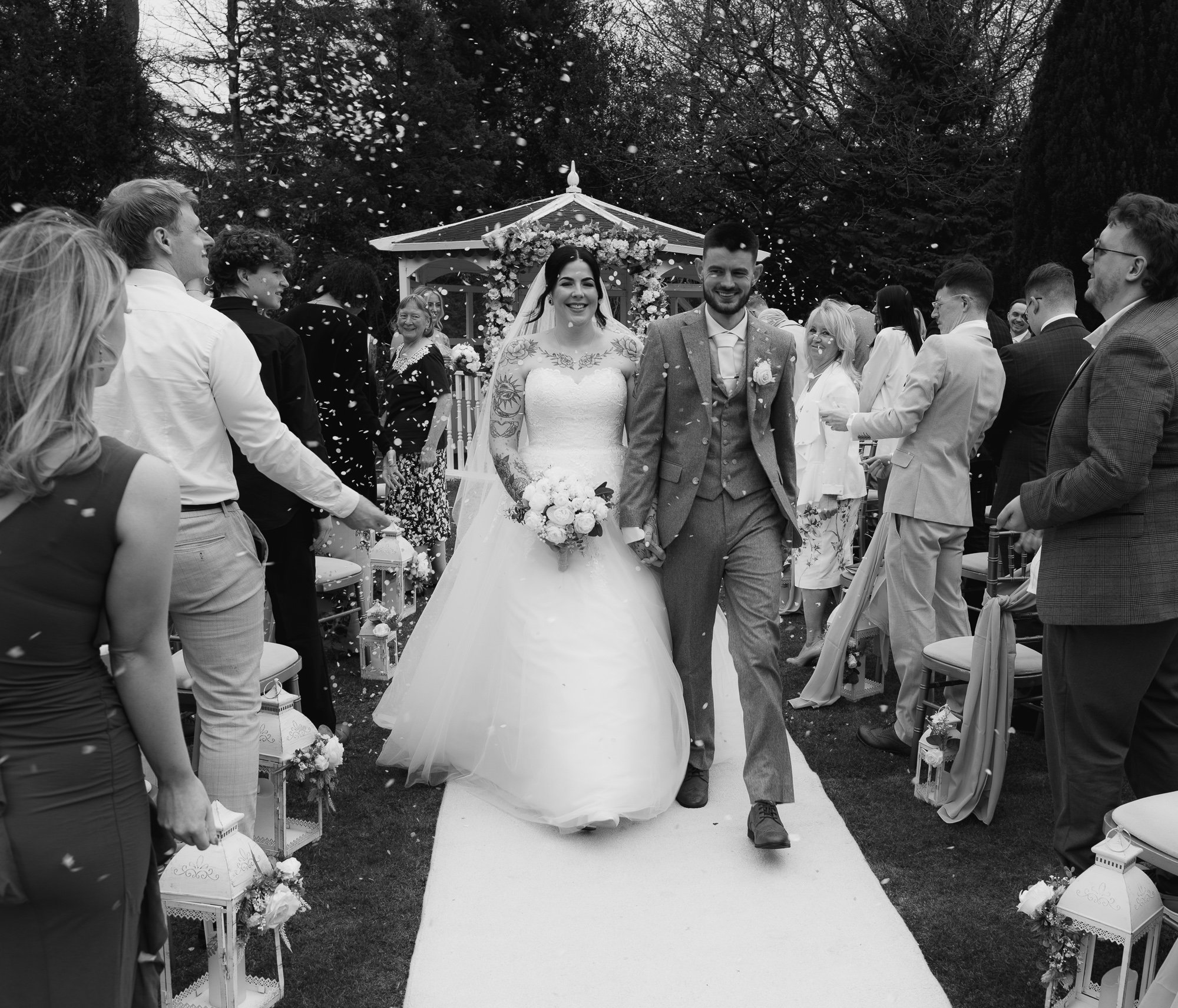 Bride and groom walking down the aisle outdoors, surrounded by guests throwing confetti.