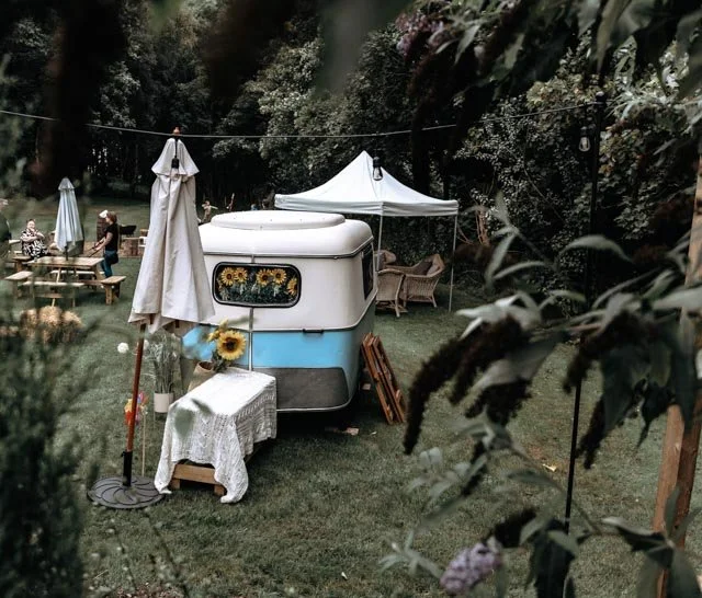 A vintage caravan set up in a garden with outdoor seating, a canopy tent, and decorated with sunflowers and umbrellas. Trees surround the area, creating a serene outdoor setting.