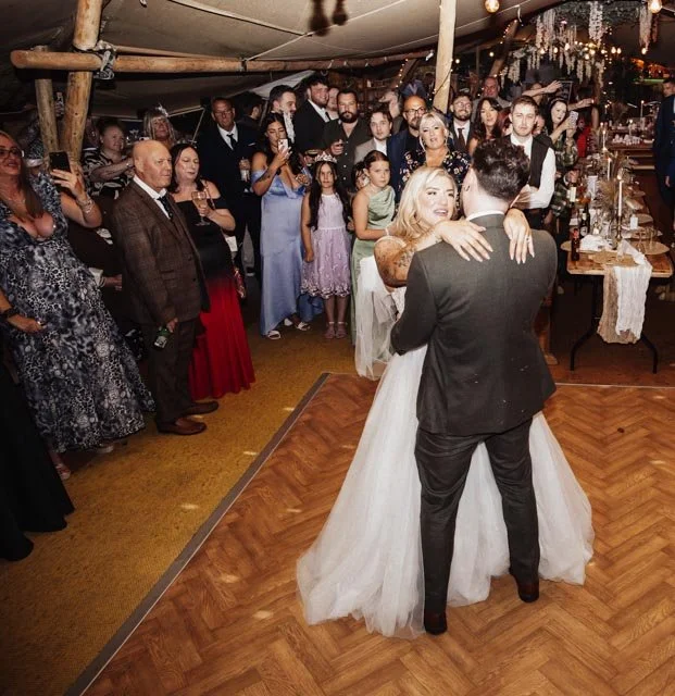 Bride and groom sharing a first dance surrounded by guests at a wedding reception inside a decorated tent.