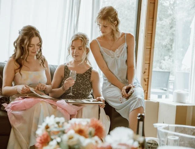 Three women sitting on a couch looking at a photo album, with one holding a champagne glass, in a cozy indoor setting with flowers on a table in the foreground.