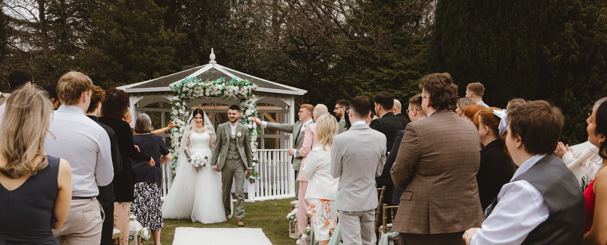 Outdoor wedding ceremony with bride and groom walking down the aisle, surrounded by applauding guests, near a floral-decorated archway and gazebo.