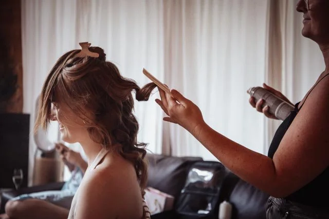 Hairstylist curling a woman's hair with a brush and hairspray at a salon.