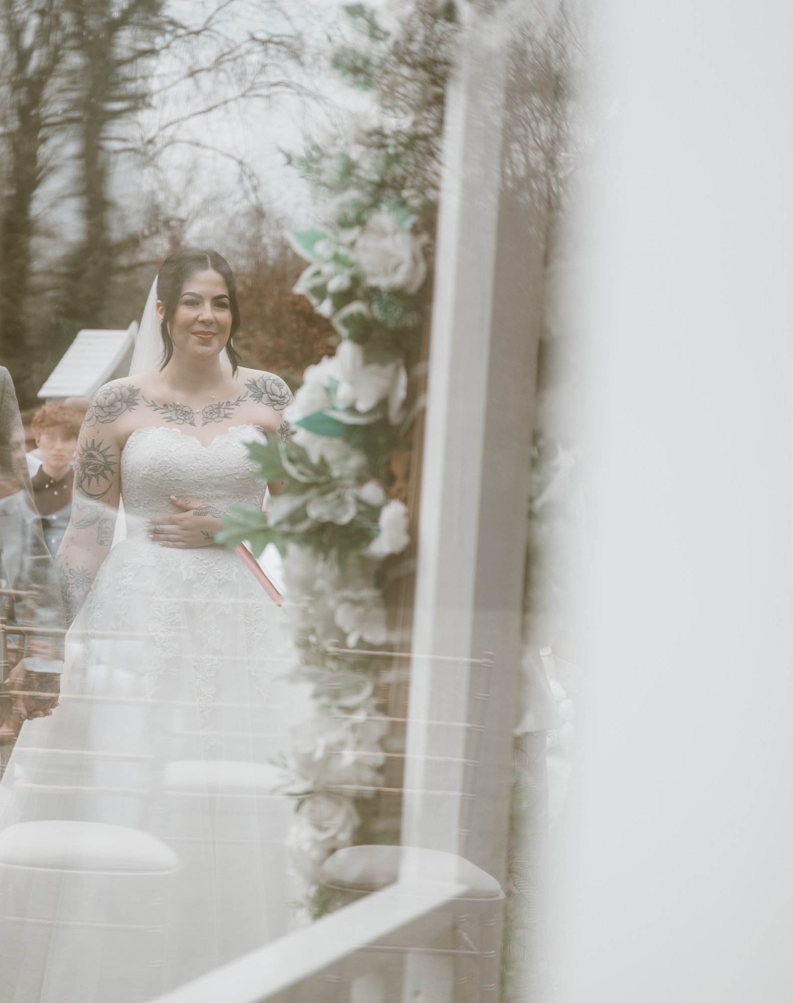 Bride in lace wedding dress with tattoos, standing near floral decorations, reflected in glass.