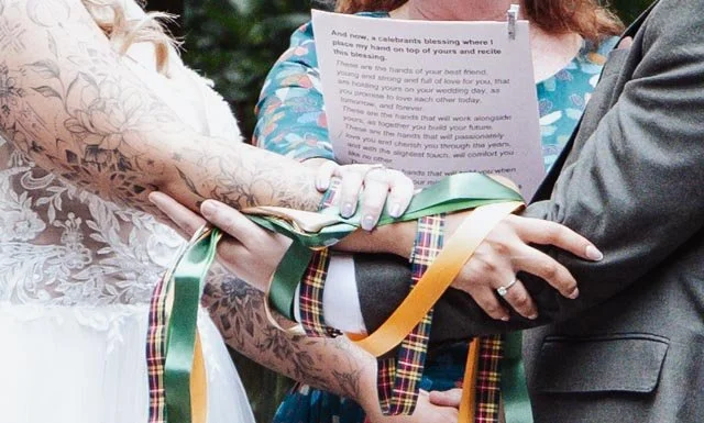 Close-up of a couple's hands tied with colorful ribbons during a wedding handfasting ceremony, with a person holding a script in the background.