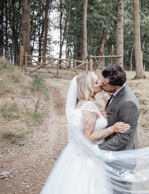 Bride and groom kissing on a rustic path with trees and a gate in the background.