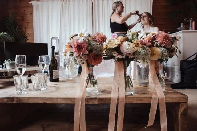 Wedding preparation scene with floral arrangements on a table, champagne bottle, and glasses. A woman styles another woman's hair in the background.