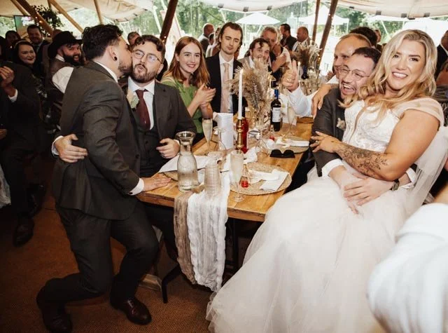 A joyful wedding celebration with a bride and groom, surrounded by guests seated at a long table under a tent. The setting is lively with people laughing and enjoying the event. The table is adorned with decorative elements and drinks.