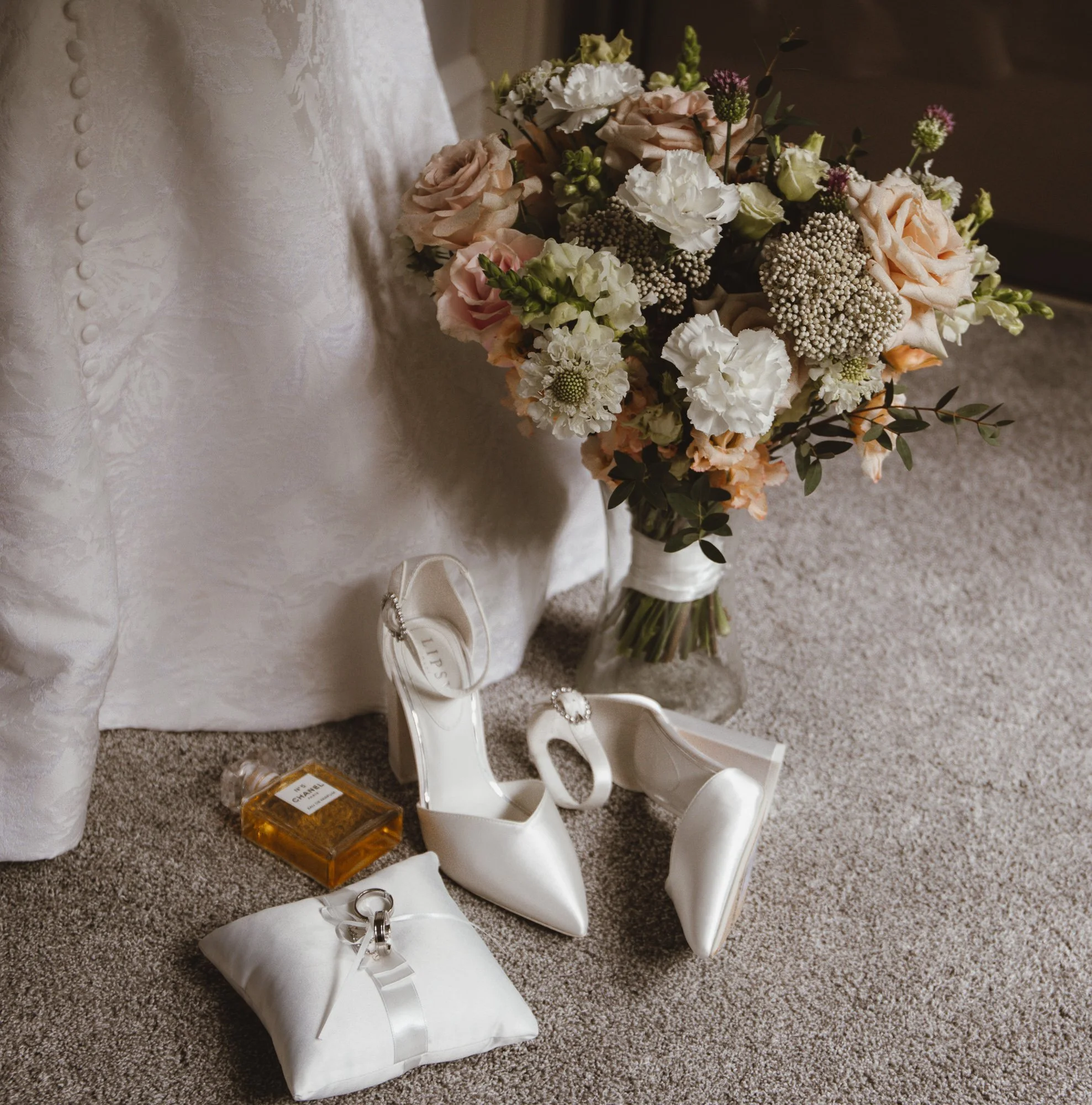 A bouquet of pink and white flowers, white high-heeled shoes, a white pillow with rings, a small perfume bottle, and an ivory satin dress fabric with buttons.