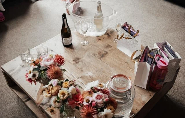 A wooden coffee table with wine bottles, glasses, and a pitcher. There are floral arrangements and gift bags on the table.