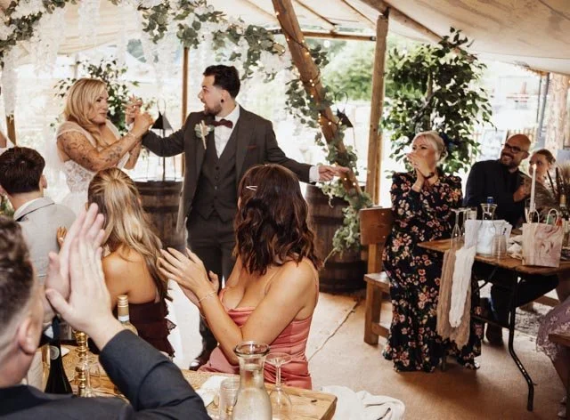 Wedding reception with guests applauding and a couple standing in the center under a tent decorated with greenery.