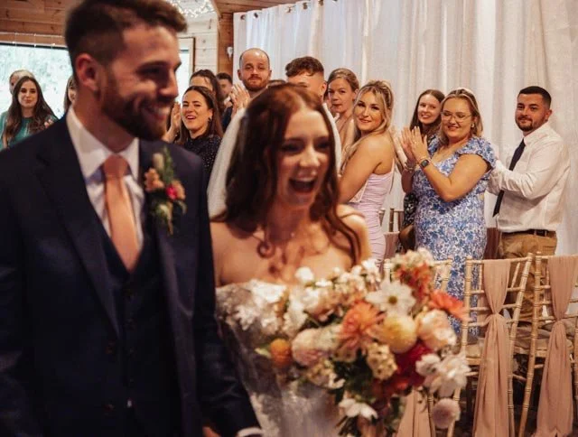 Wedding ceremony with happy bride and groom walking down the aisle as guests applaud, bride holding bouquet of flowers, decorated venue with wooden interior and chairs.