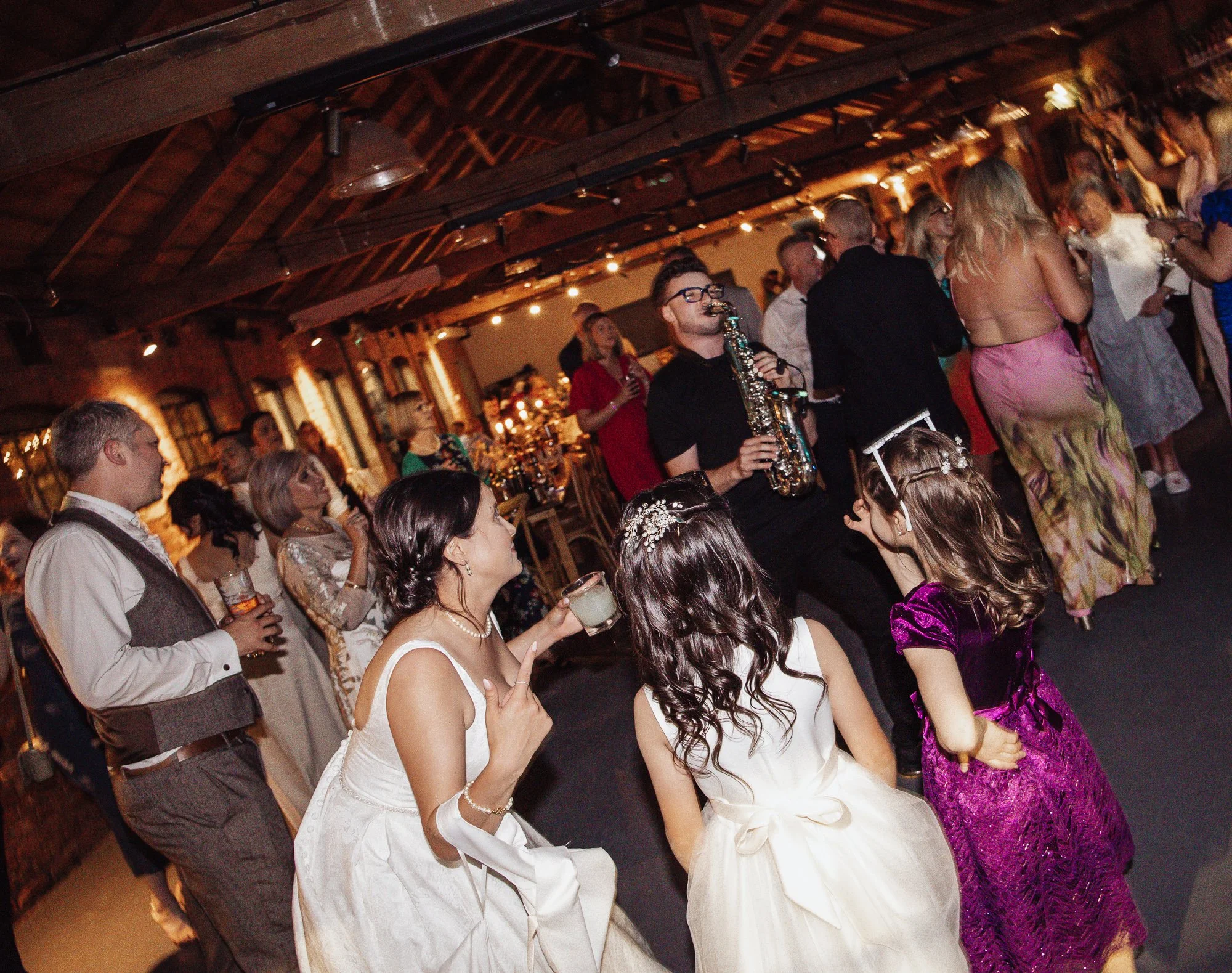 People dancing and socializing at a celebration in a rustic event space with exposed wooden beams, including women in elegant dresses and a musician playing the saxophone.