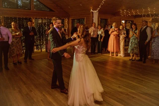 A couple dancing at a wedding reception in a warmly-lit wooden hall, surrounded by guests watching and celebrating with string lights in the background.