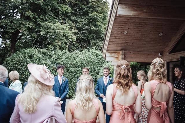 Group of people in formal attire at an outdoor event, with women wearing pink dresses and men in blue suits, surrounded by greenery and a wooden building.