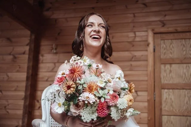 Bride holding a colorful bouquet of flowers, smiling joyfully, in a wooden interior setting.