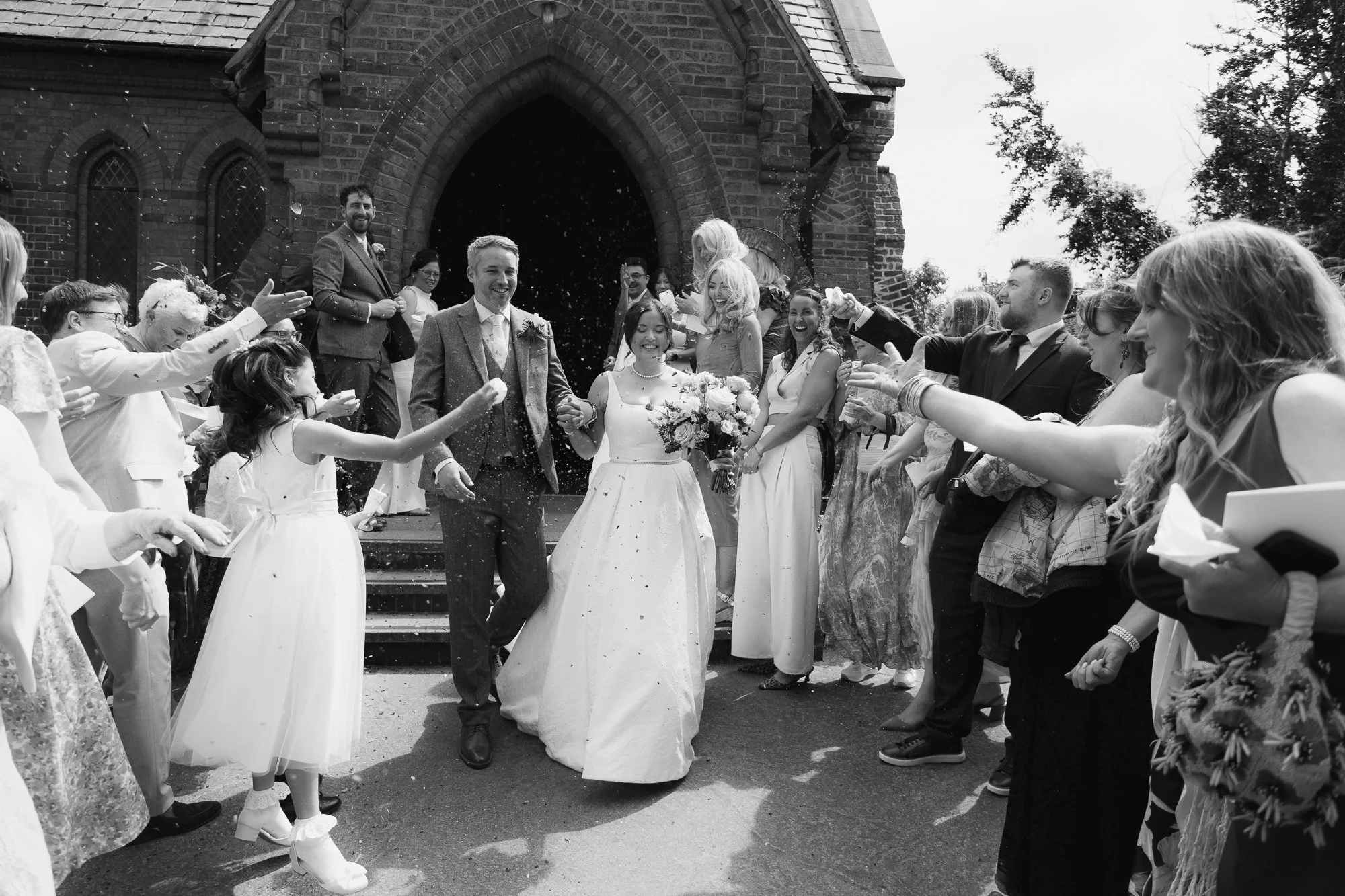 A black and white photo of a wedding celebration with the bride and groom leaving a church and being showered with confetti by friends and family.