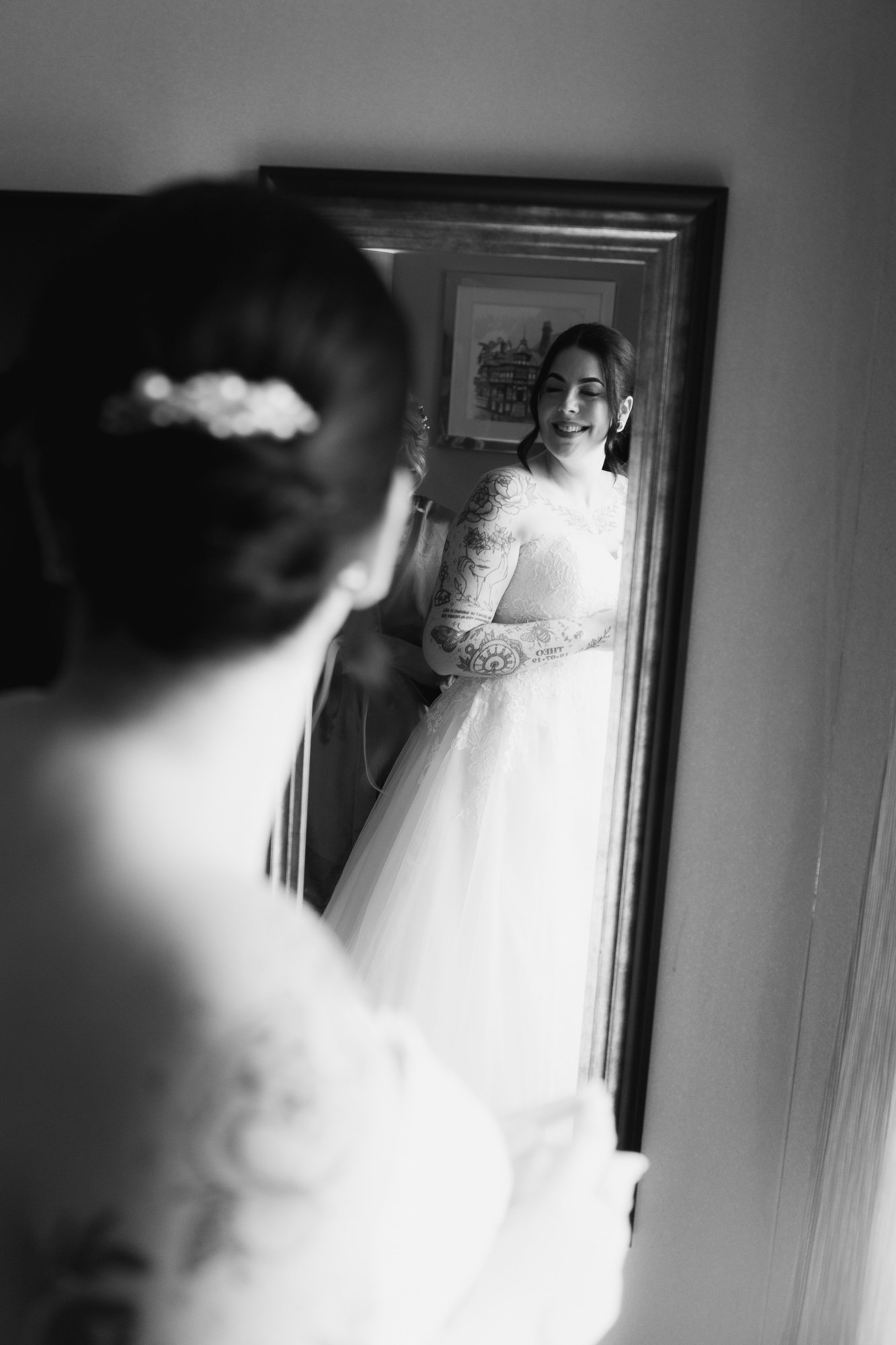 Bride smiling at her reflection in a mirror, wearing a wedding dress with tattooed sleeves, captured in black and white.