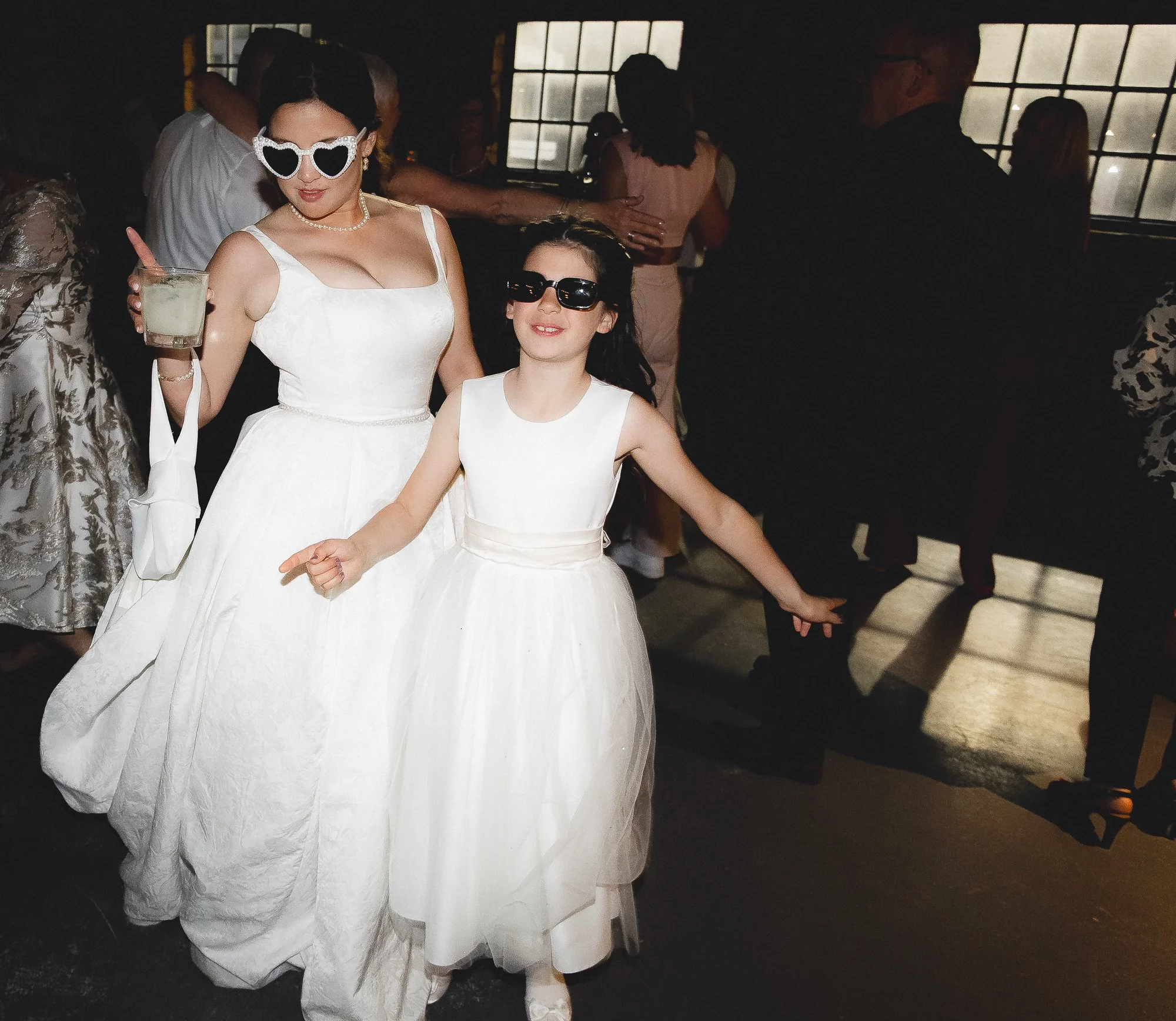 Two women dressed in white at a party or wedding, wearing sunglasses, and dancing with other guests in the background.