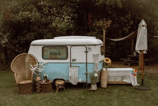 Vintage camper van setup with bohemian decor including wicker furniture, sunflower, and plants in a grassy area surrounded by trees.
