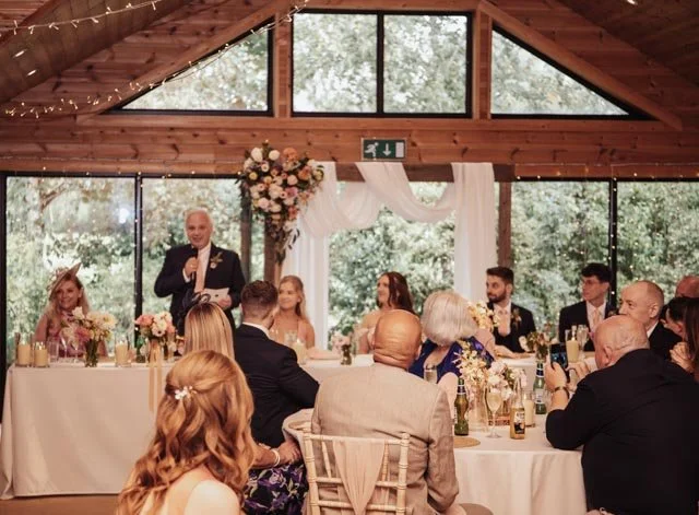 A wedding reception with a man giving a speech at a long table. Guests are seated around, listening and holding drinks. The room is decorated with flowers and fairy lights, and large windows show greenery outside. People are dressed formally.