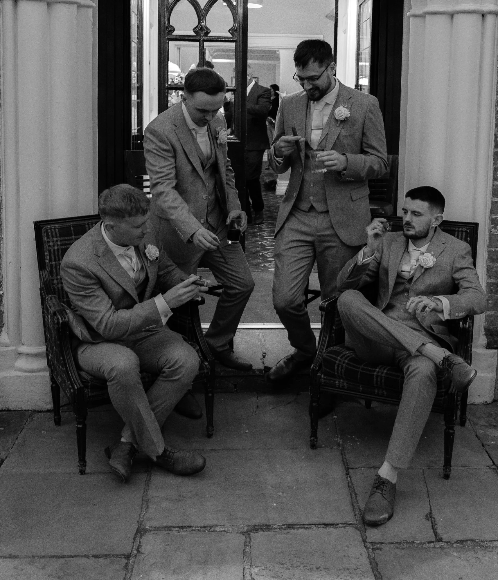 Four men in formal suits with boutonnieres, sitting and standing outside a building, engaged in conversation and holding drinks and cigars.