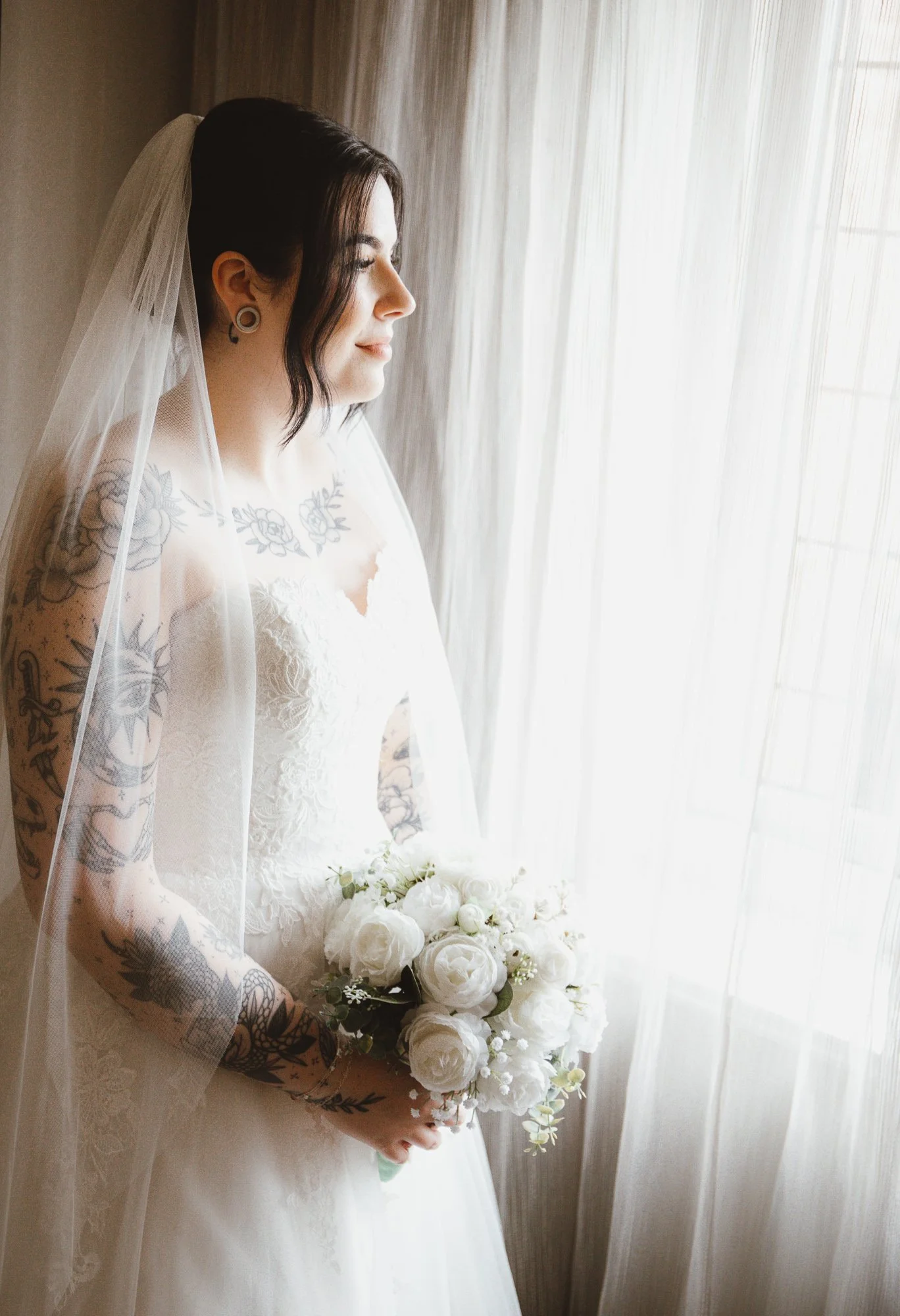 Bride in a wedding dress holding a bouquet, standing by a window, with tattoos on her arms, and wearing a veil.
